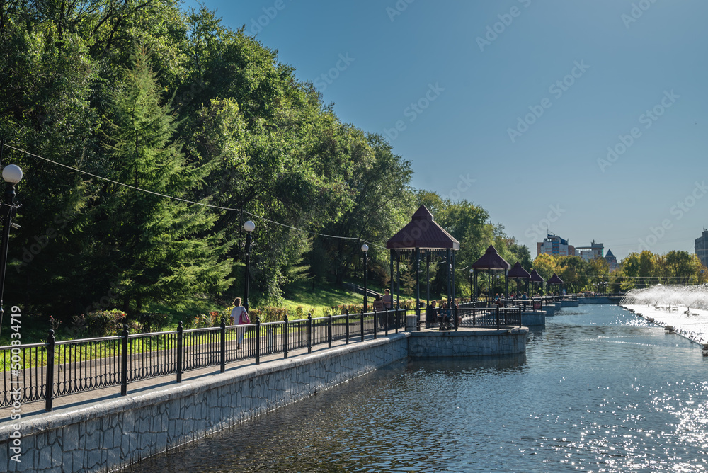 Fototapeta premium A beautiful autumn view of the public park with the pond, trees, fountains, and people walking in distance