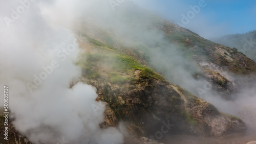 Clouds of steam from hot springs envelop the mountain slopes. Green vegetation on the hills. Blue sky. Poor visibility due to haze. Kamchatka. Valley of Geysers