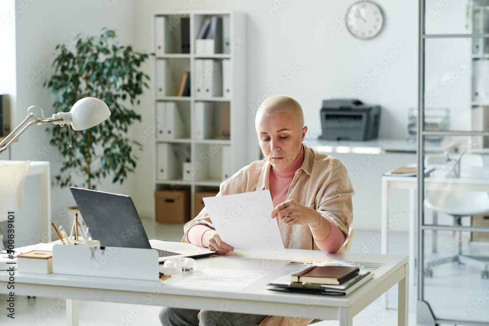 Serious young female analyst in casual outfit sitting at desk with laptop and reading business paper in office Serious young female analyst in casual outfit sitting at desk with laptop and reading business paper in office