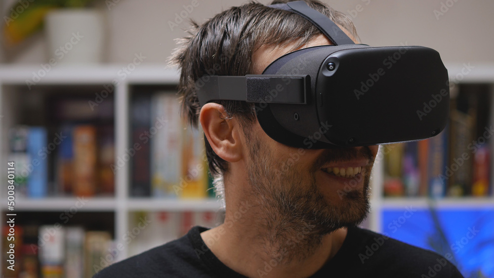 A bearded man wearing VR glasses smiles at the virtual world he sees. Close-up. In the background is a rack of games. VR helmet for entertainment, education, and gaming. A bearded man wearing VR glasses smiles at the virtual world he sees. Close-up. In the background is a rack of games. VR helmet for entertainment, education, and gaming.