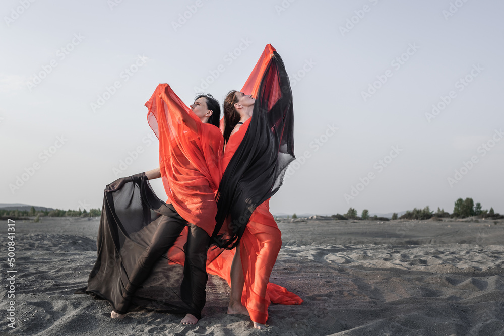 Two girls in black and red flowing fabric pose on sand dune Two girls in black and red flowing fabric pose on sand dune