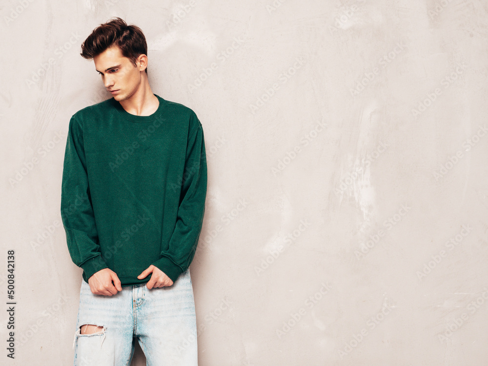 Portrait of handsome confident stylish hipster lambersexual model.Man dressed in green sweater and jeans. Fashion male posing in studio near grey wall