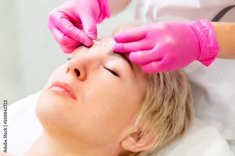Fototapeta premium Mesotherapy and rejuvenation. Portrait of an adult blonde woman at a cosmetologist's appointment. A beautician in pink medical gloves applies a needle to the client's face