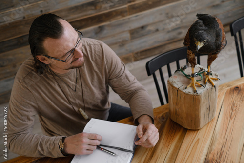 Man is working, writing with wild bird at home by the table. Making noted, memories, diary with eagle as pet. Unusual animals at home. Human friendship. Taking care of buzzard Top iew