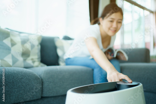 Happy Asian young woman turning on high efficiency air purifier while staying and reading a book in the living room. 