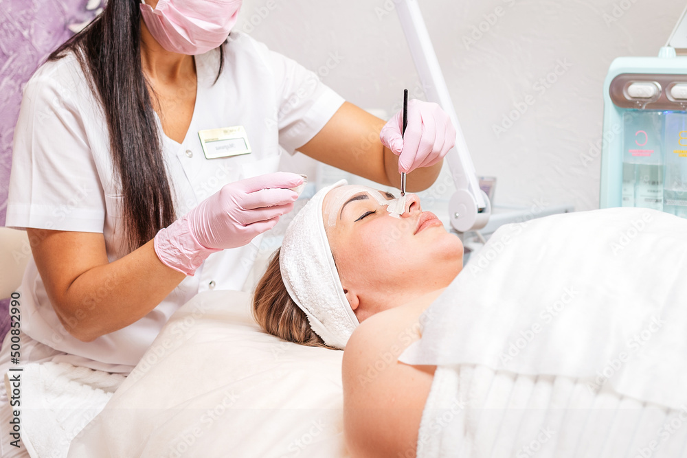 A cosmetologist in a medical mask and gloves holds a bowl of cosmetics, applying a brush mask on the client's face. Side view. Copy space. Concept of professional cosmetology during coronavirus