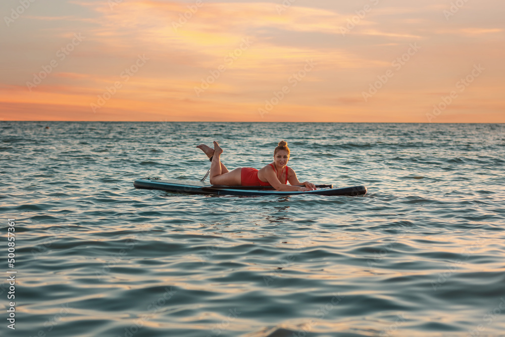 Plus sized pretty woman in red swimsuit posing lying on a sup board. Copy space. Sunset sky on the background. The concept of water sports and summer vacation