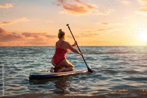Plus sized beautiful Caucasian woman in red swimsuit swimming sitting on a sup board. Sunset on the background. Back view. The concept of sports and summer vacation