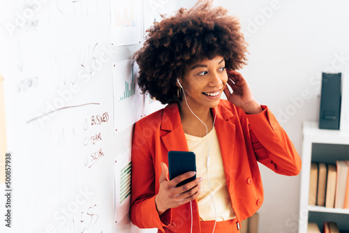 Smiling businesswoman holding smart phone listening music through in-ear headphones at office photo