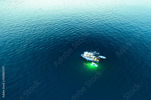 Fototapeta Naklejka Na Ścianę i Meble -  Fishing boat illuminates water with green flashlight light at fishing and drifting on sea surface. Calm Adriatic sea reflecting sunset at twilight. Aerial view
