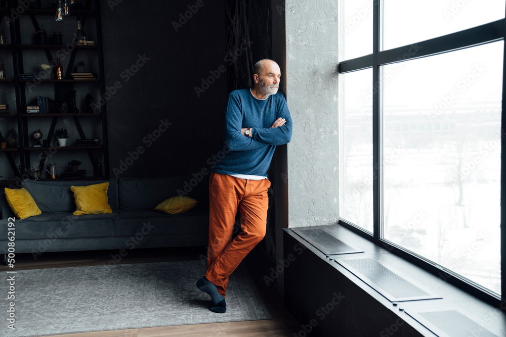 Senior man standing with arms crossed looking through window at home Senior man standing with arms crossed looking through window at home