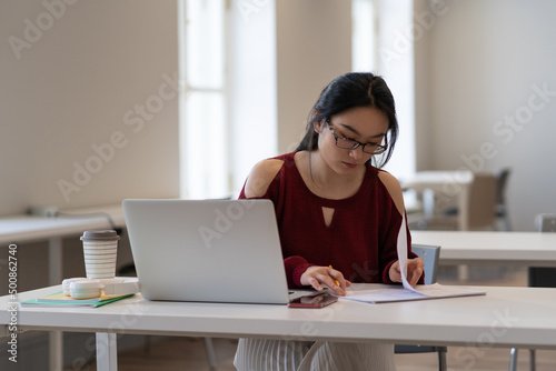 Young student girl making homework in library. Asian female writing notes for educational project in university or college preparing for course work, test or graduation exam using laptop at campus