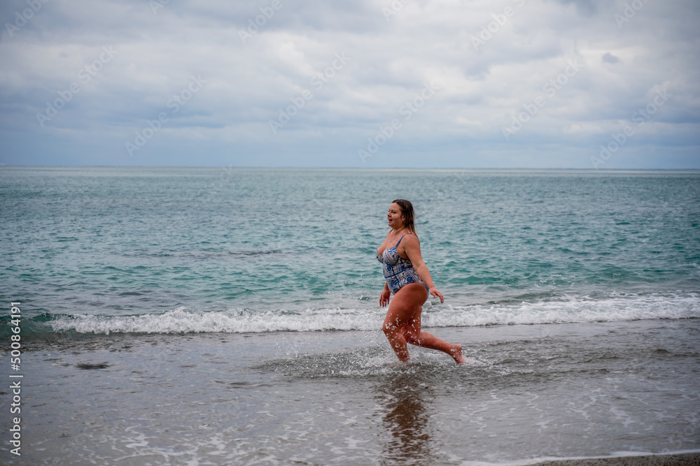 A plump woman in a bathing suit enters the water during the surf. Alone on the beach, Gray sky in the clouds, swimming in winter. A plump woman in a bathing suit enters the water during the surf. Alone on the beach, Gray sky in the clouds, swimming in winter.