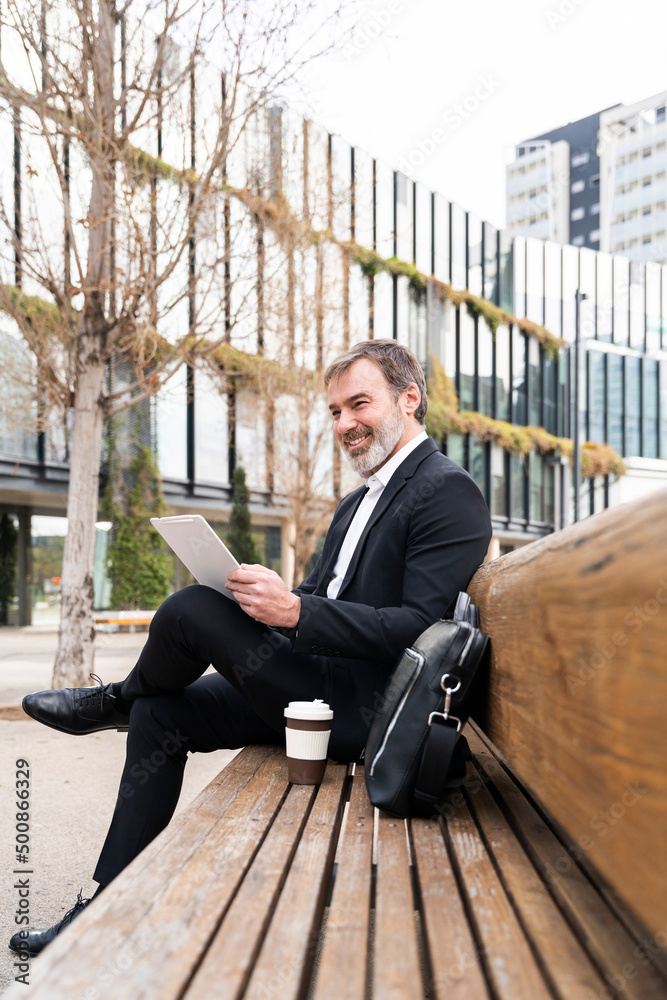 Smiling businessman with tablet PC sitting by laptop bag on bench