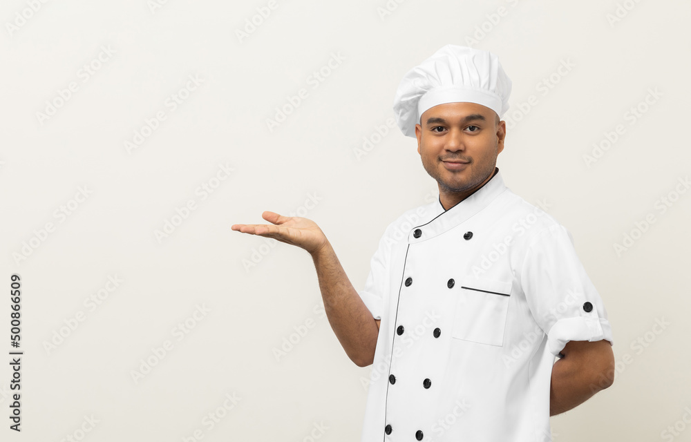 Smiling Young handsome asian man chef in uniform with hat standing showing hand palm up holding some menu on isolated. Cooking indian man Occupation chef People in kitchen restaurant and hotel. Smiling Young handsome asian man chef in uniform with hat standing showing hand palm up holding some menu on isolated. Cooking indian man Occupation chef People in kitchen restaurant and hotel.