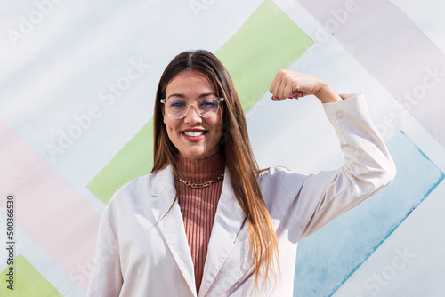 Smiling woman flexing muscles standing in front of wall on sunny day