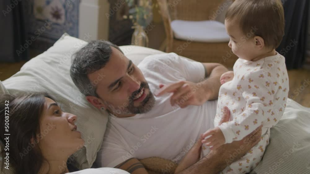 Happy Caucasian couple lying in bed on Sunday morning and playing with their cute baby girl. Bearded mature man holding his adorable infant, then baby kissing his cheek. Family, parenthood concept Happy Caucasian couple lying in bed on Sunday morning and playing with their cute baby girl. Bearded mature man holding his adorable infant, then baby kissing his cheek. Family, parenthood concept