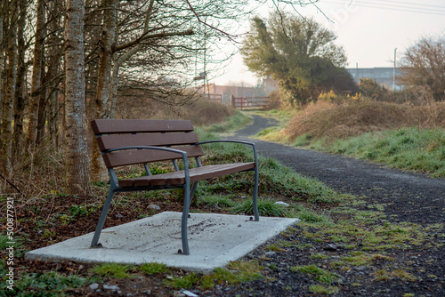 Brand new brown bench with metal frame and plastic parts installed on a concrete base by a small footpath in a park. Solid and practical design. Nobody