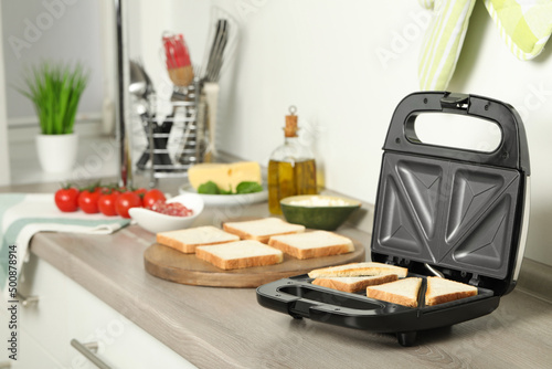 Fotografia Modern sandwich maker with bread slices on wooden table in kitchen