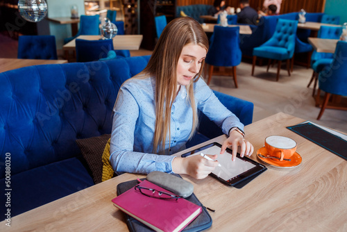 Young woman is working on her tablet in the cafe, cup of coffee next to her on the table. 