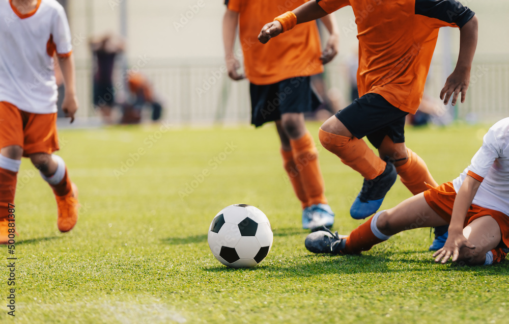 Group of multi-ethnic children playing soccer game. Young boys running after soccer ball on grass football field. Kids in orange and white jersey shirts. The player try to tackle a soccer ball