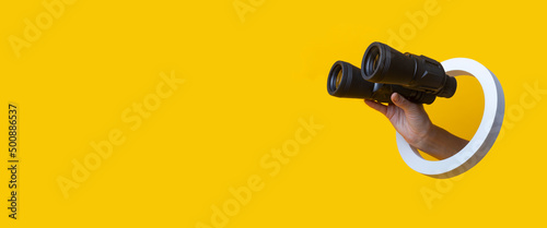 Female hand holds binoculars through a hole on a yellow background. Banner.