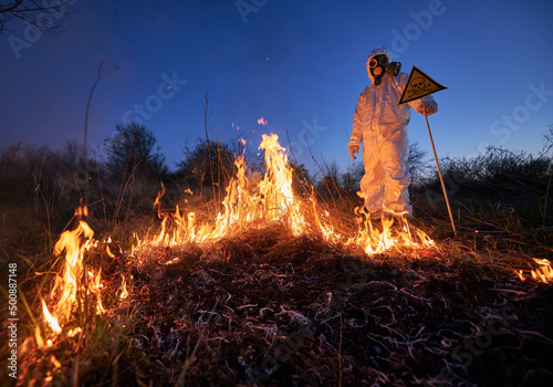 Firefighter ecologist fighting fire in field at night. Man in protective suit and gas mask near burning grass with smoke, holding warning sign with skull and crossbones. Natural disaster concept.