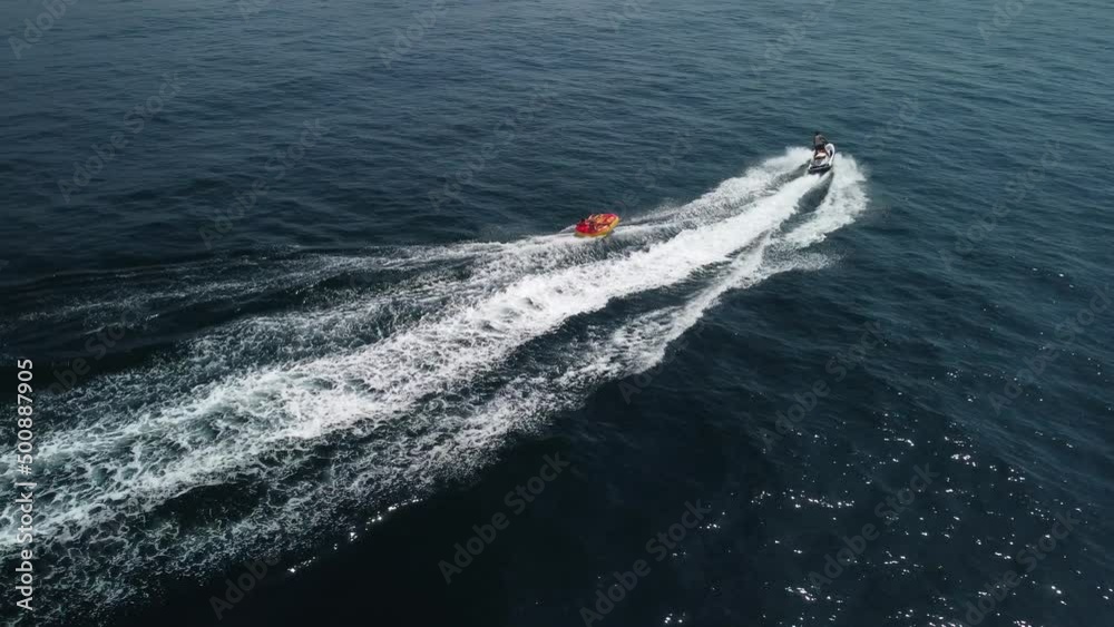 Happy people are going to swim on an air mattress behind a jet ski. Tourists ride the inflatable watercraft boat. Sea attraction. Jet ski skating people on a large Inflatable raft. Aerial copter view. Happy people are going to swim on an air mattress behind a jet ski. Tourists ride the inflatable watercraft boat. Sea attraction. Jet ski skating people on a large Inflatable raft. Aerial copter view.