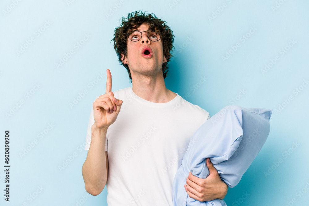 Young caucasian man wearing a pajama and holding pillow isolated on blue background pointing upside with opened mouth. Young caucasian man wearing a pajama and holding pillow isolated on blue background pointing upside with opened mouth.