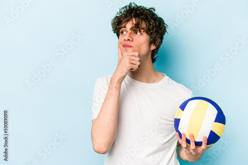 Young caucasian man playing volleyball isolated on blue background looking sideways with doubtful and skeptical expression.