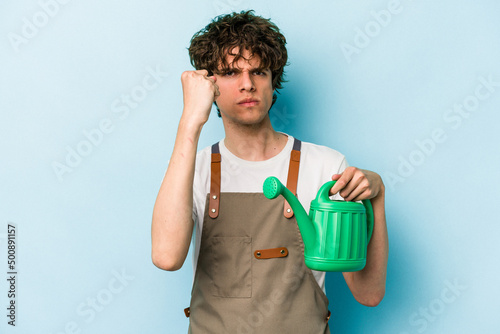 Young gardener caucasian man holding watering can isolated on blue background showing fist to camera, aggressive facial expression.