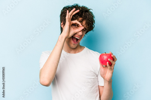 Young caucasian man holding an apple isolated on blue background excited keeping ok gesture on eye.