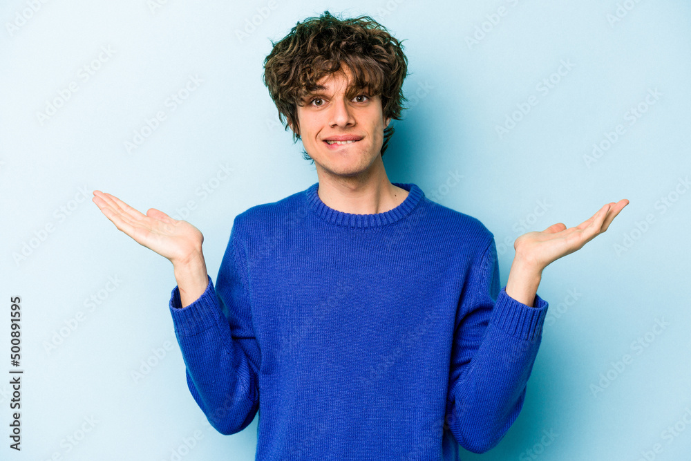 Young caucasian man isolated on blue background confused and doubtful shrugging shoulders to hold a copy space.