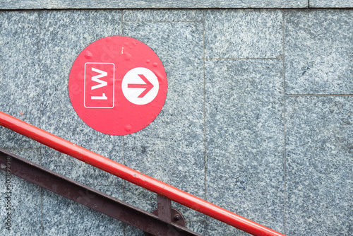 Underground indication on a red circle with white arrow on the wall of Milan underground, Lombardy, Italy. Red diagonal handrail in the foreground. The writing 