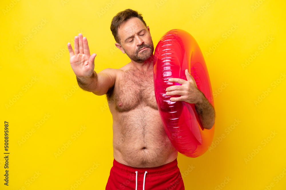 Middle age caucasian man holding inflatable donut isolated on yellow background making stop gesture and disappointed