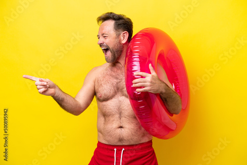 Middle age caucasian man holding inflatable donut isolated on yellow background pointing finger to the side and presenting a product © luismolinero