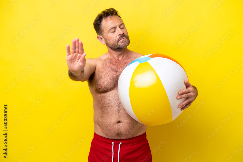 Middle age caucasian man holding beach ball isolated on yellow background making stop gesture and disappointed