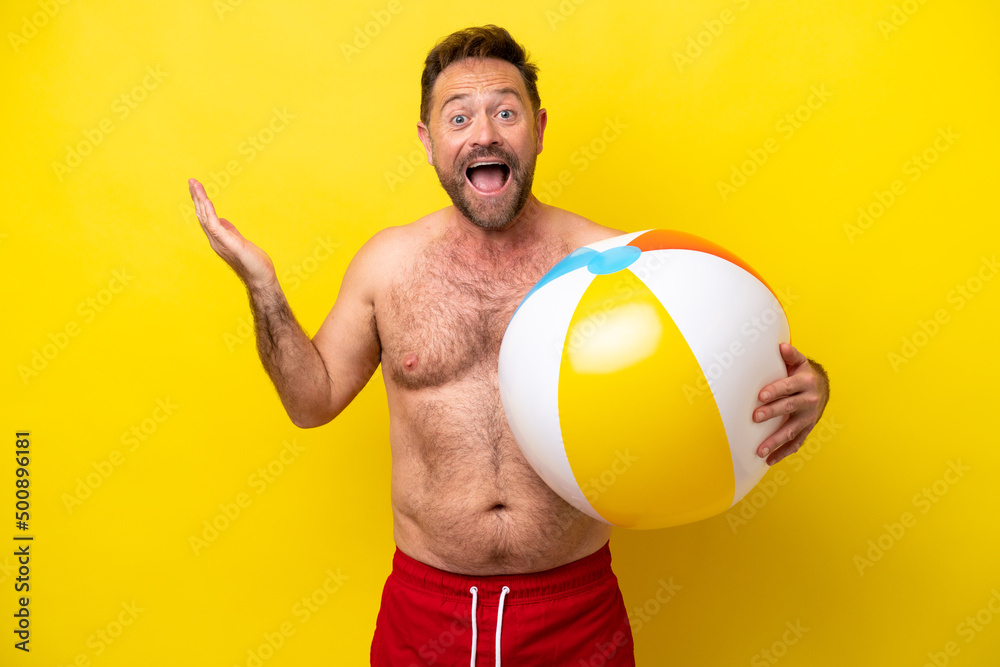 Middle age caucasian man holding beach ball isolated on yellow background with shocked facial expression