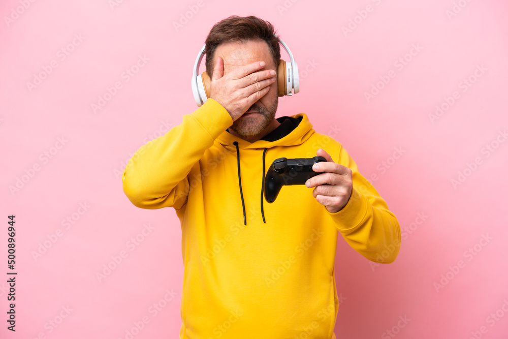 Middle age man playing with a video game controller isolated on pink background covering eyes by hands. Do not want to see something Middle age man playing with a video game controller isolated on pink background covering eyes by hands. Do not want to see something