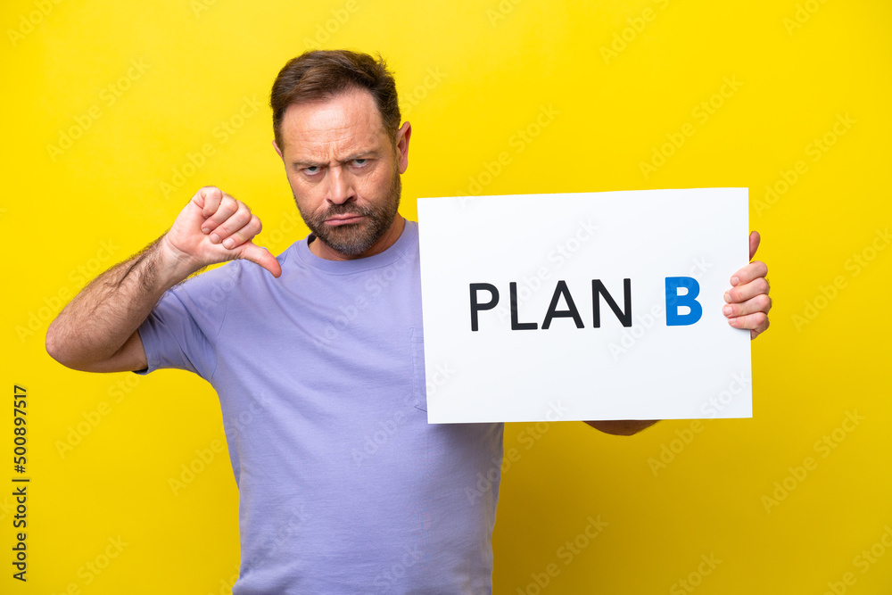 Middle age caucasian man isolated on yellow background holding a placard with the message PLAN B and doing bad signal Middle age caucasian man isolated on yellow background holding a placard with the message PLAN B and doing bad signal