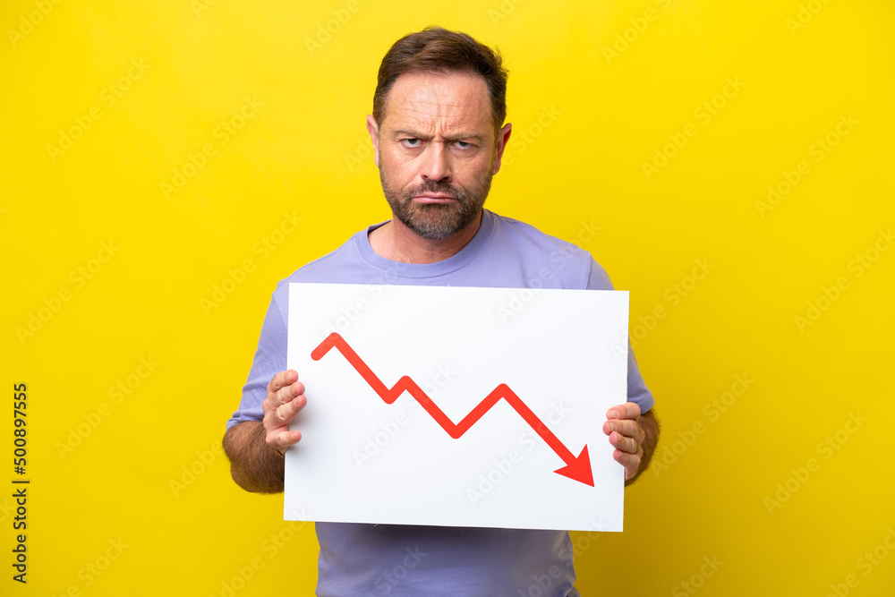 Middle age caucasian man isolated on yellow background holding a sign with a decreasing statistics arrow symbol with sad expression Middle age caucasian man isolated on yellow background holding a sign with a decreasing statistics arrow symbol with sad expression