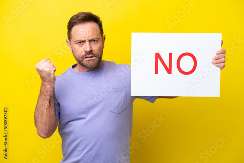 Middle age caucasian man isolated on yellow background holding a placard with text NO and angry © luismolinero