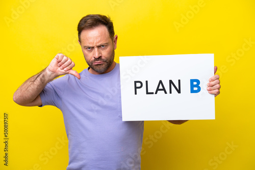 Middle age caucasian man isolated on yellow background holding a placard with the message PLAN B and doing bad signal © luismolinero