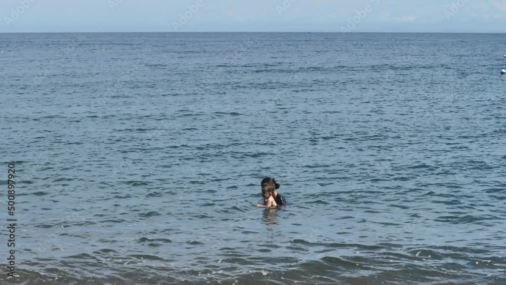 At a far distance, a young mother with a small child is shown, who spend their free leisure swimming in the sea, strengthening their health. The weather is calm. Baby is 9 months old. At a far distance, a young mother with a small child is shown, who spend their free leisure swimming in the sea, strengthening their health. The weather is calm. Baby is 9 months old.