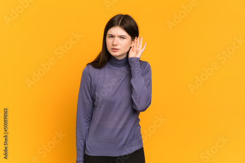 Young Ukrainian girl isolated on yellow background listening to something by putting hand on the ear