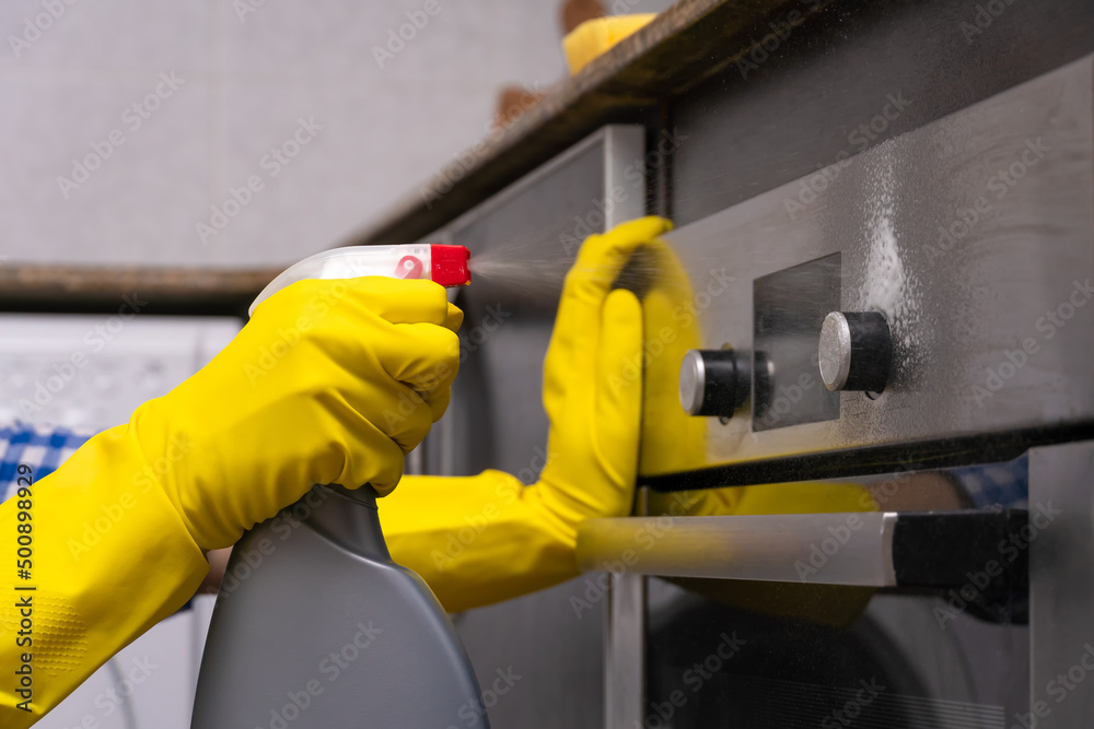 Women's hands in yellow rubber gloves are rubbing cleaning agent on the surface of the oven in the home kitchen. Concept of housework and housekeeping Women's hands in yellow rubber gloves are rubbing cleaning agent on the surface of the oven in the home kitchen. Concept of housework and housekeeping