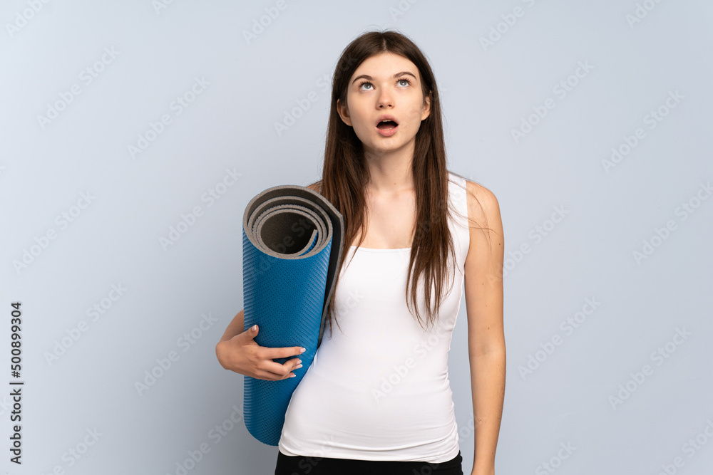 Young Ukrainian sport girl going to yoga classes while holding a mat looking up and with surprised expression Young Ukrainian sport girl going to yoga classes while holding a mat looking up and with surprised expression