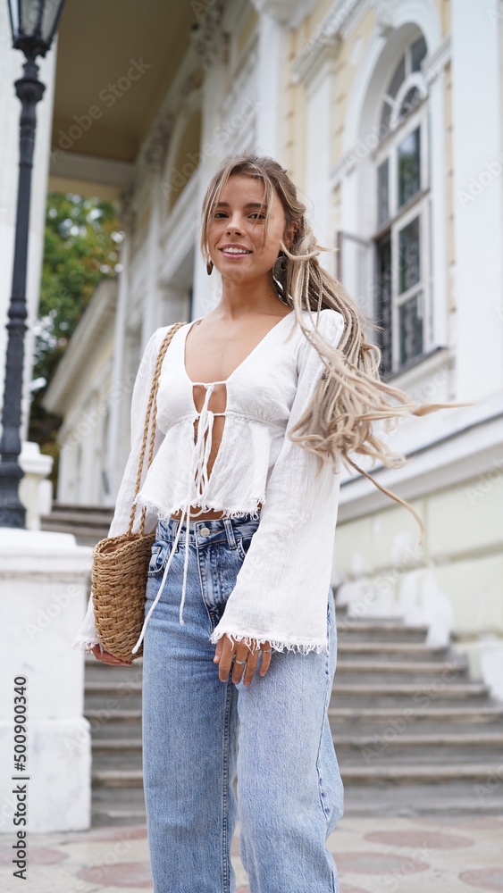 Fototapeta premium Young sexy beautiful woman with blonde dreadlocks walking on the street. Close up portrait bohemian girl in white blouse and jewels hippie style. Smiling girls portrait. Traveling 