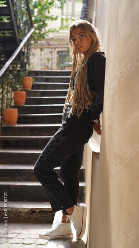 Fashionable pretty hippie woman with unusual dreads hairstyle, posing at city cafe, enjoying, toned colors. Young beautiful bohemian woman with blonde afro hairstyle dreads walking on the street . 