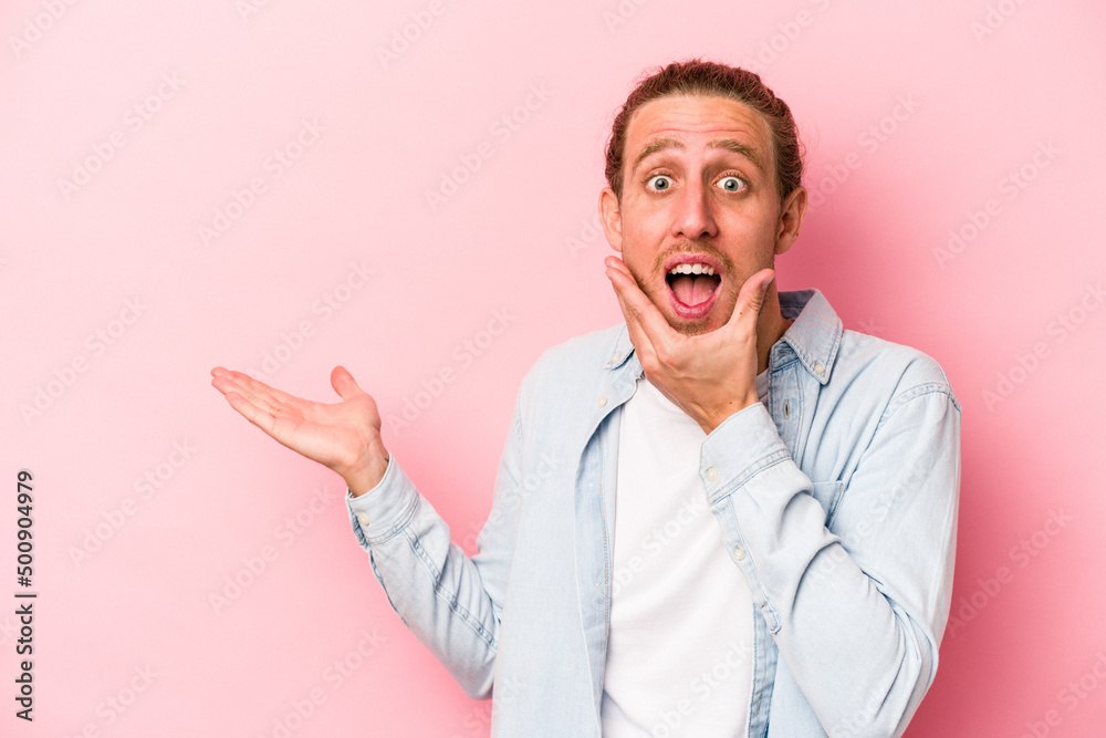 Young caucasian man isolated on pink background holds copy space on a palm, keep hand over cheek. Amazed and delighted. Young caucasian man isolated on pink background holds copy space on a palm, keep hand over cheek. Amazed and delighted.